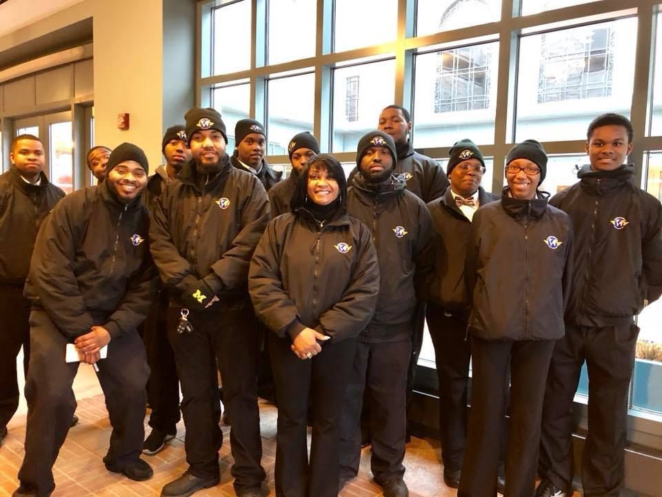 A group of people in black jackets and hats, posing indoors near a large window.