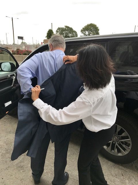 A person in a white shirt helps a man in a blue shirt and suit jacket exit a black SUV.
