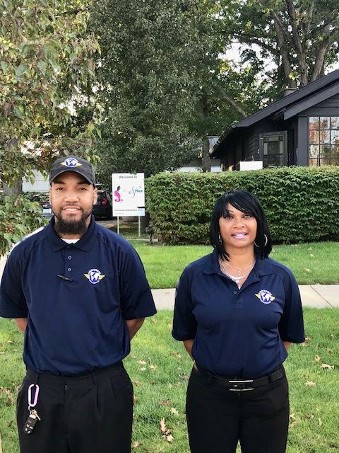 Two people in blue shirts, outdoors, smile in front of a building, green grass, and trees.