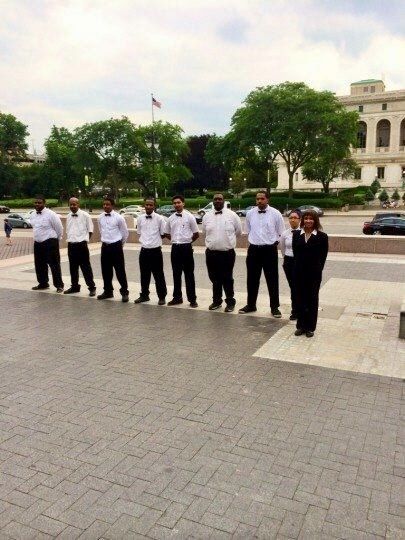 Group of people standing in front of a building, some holding hands. Gray pavement, green trees and a cloudy sky.