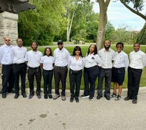 Group of people, wearing white shirts and black pants, standing outside in a park.