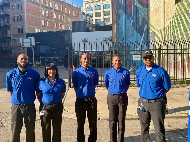 Five people in blue shirts and black pants stand in front of a fence. Buildings and a mural are in the background.