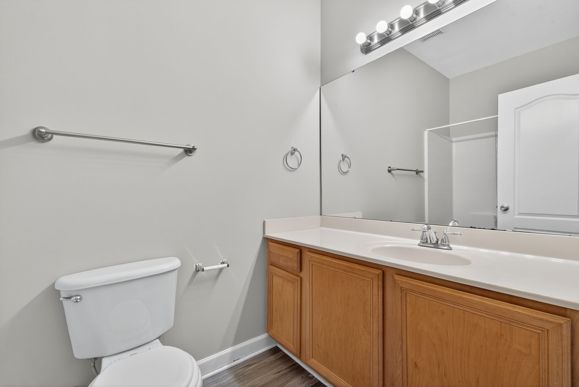 A brightly lit bathroom with a white toilet, wooden vanity, large wall mirror, and neutral grey walls.