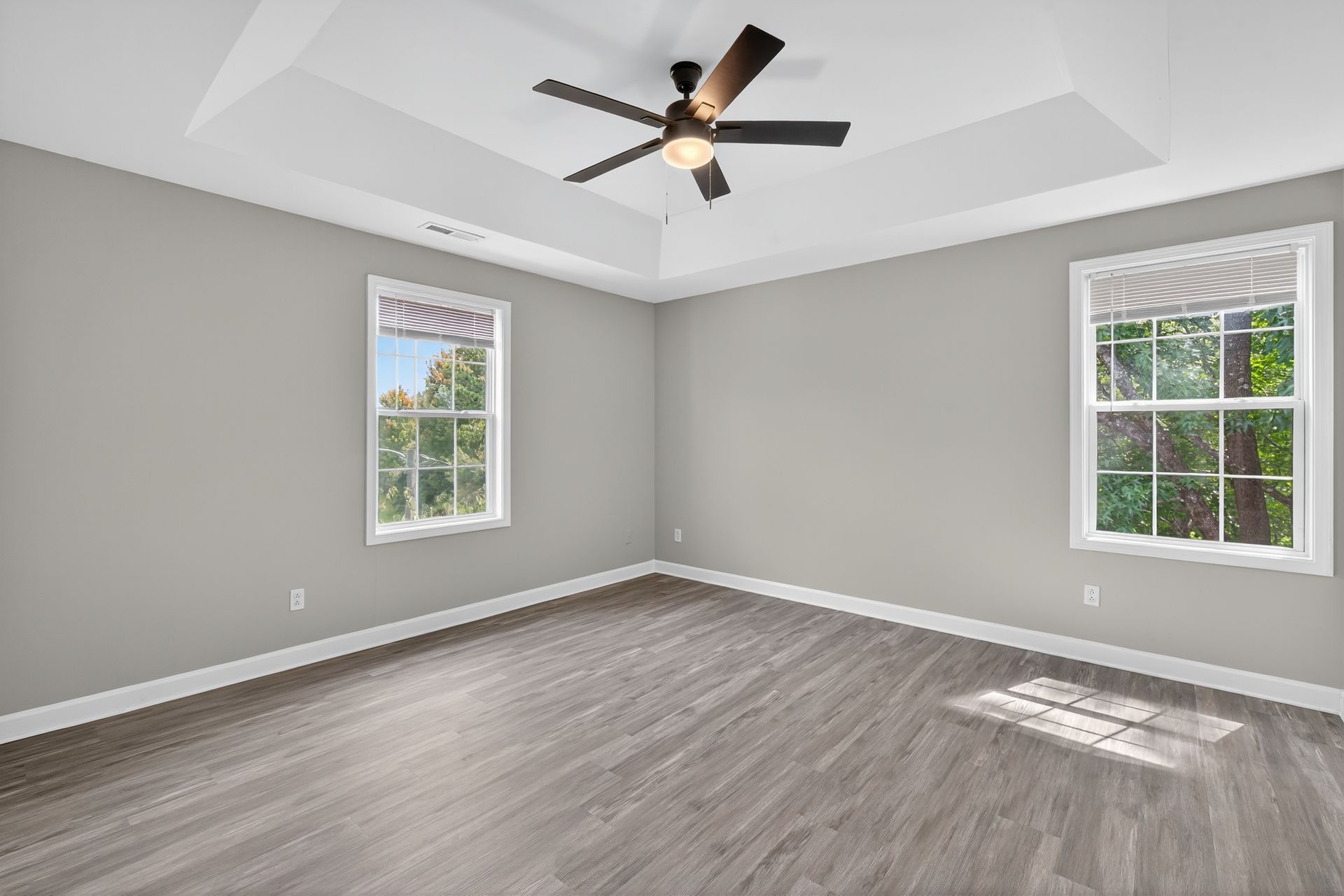Empty bedroom with gray walls, wood-look flooring, a tray ceiling with a fan, and two windows.