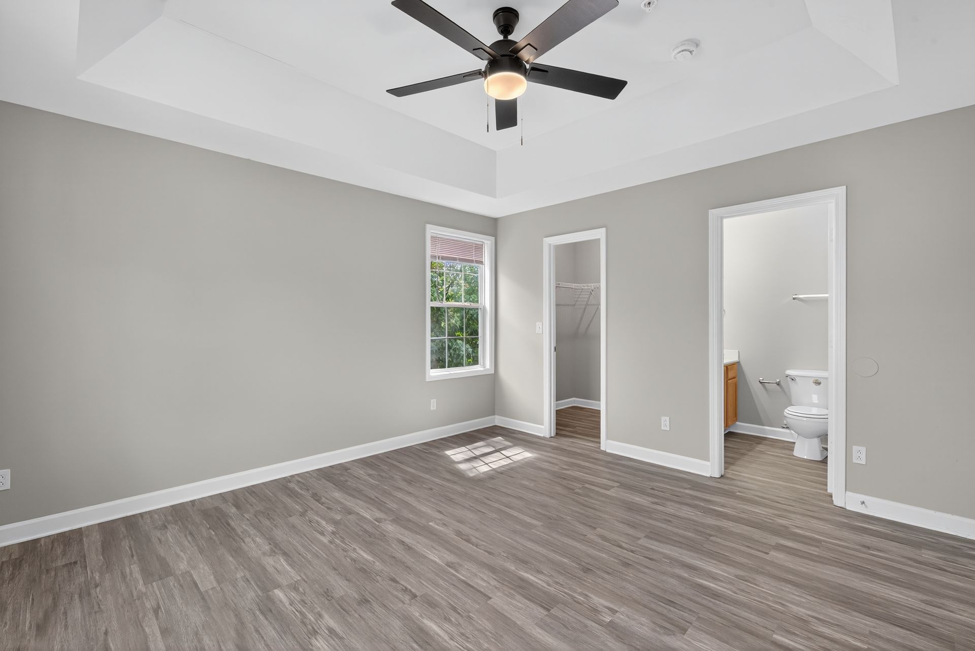 A modern bedroom with gray walls, wood-look flooring, a tray ceiling with a fan, a window, and two open doorways.