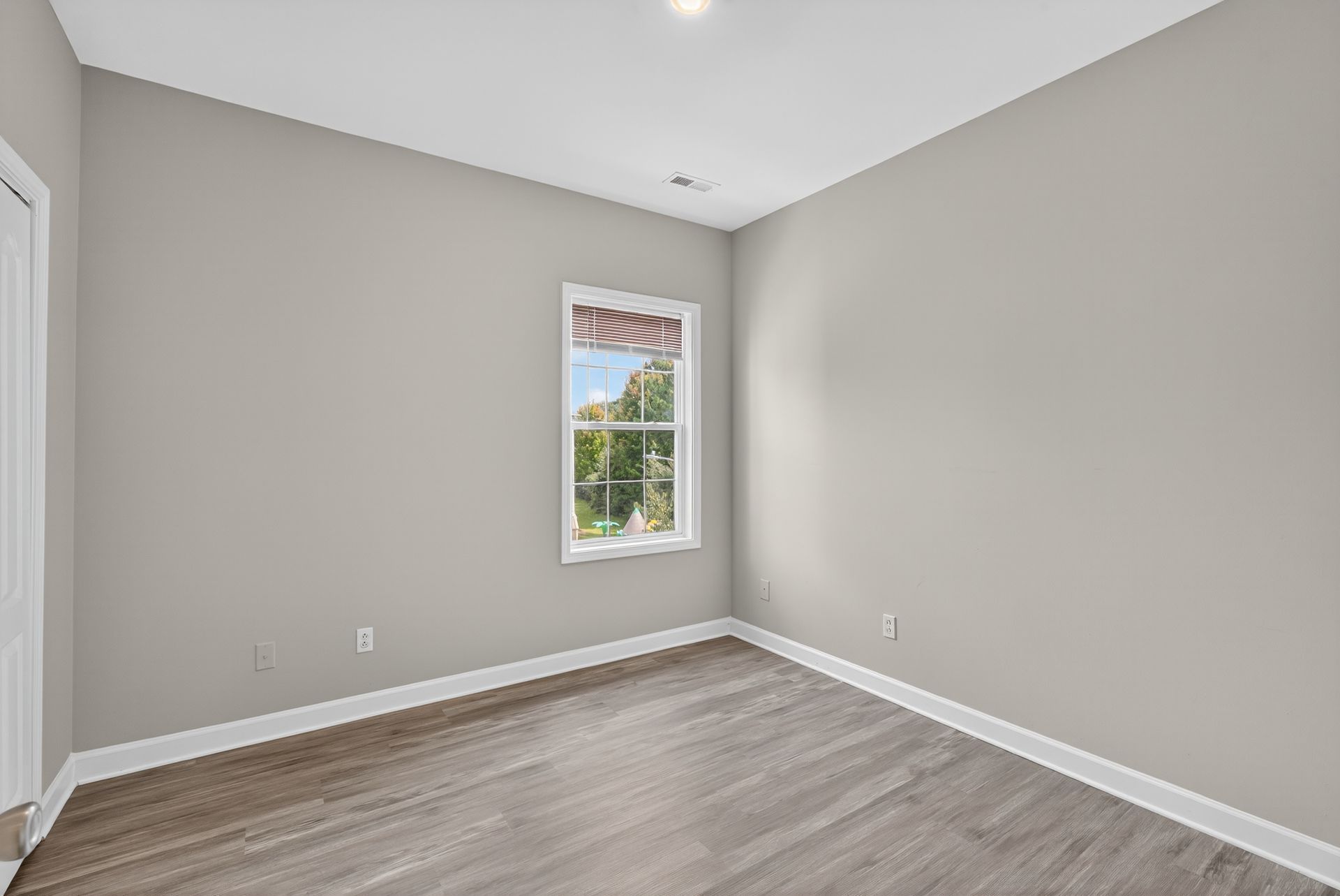 An empty bedroom with light gray walls, light wood-look flooring, a white window, and white trim.