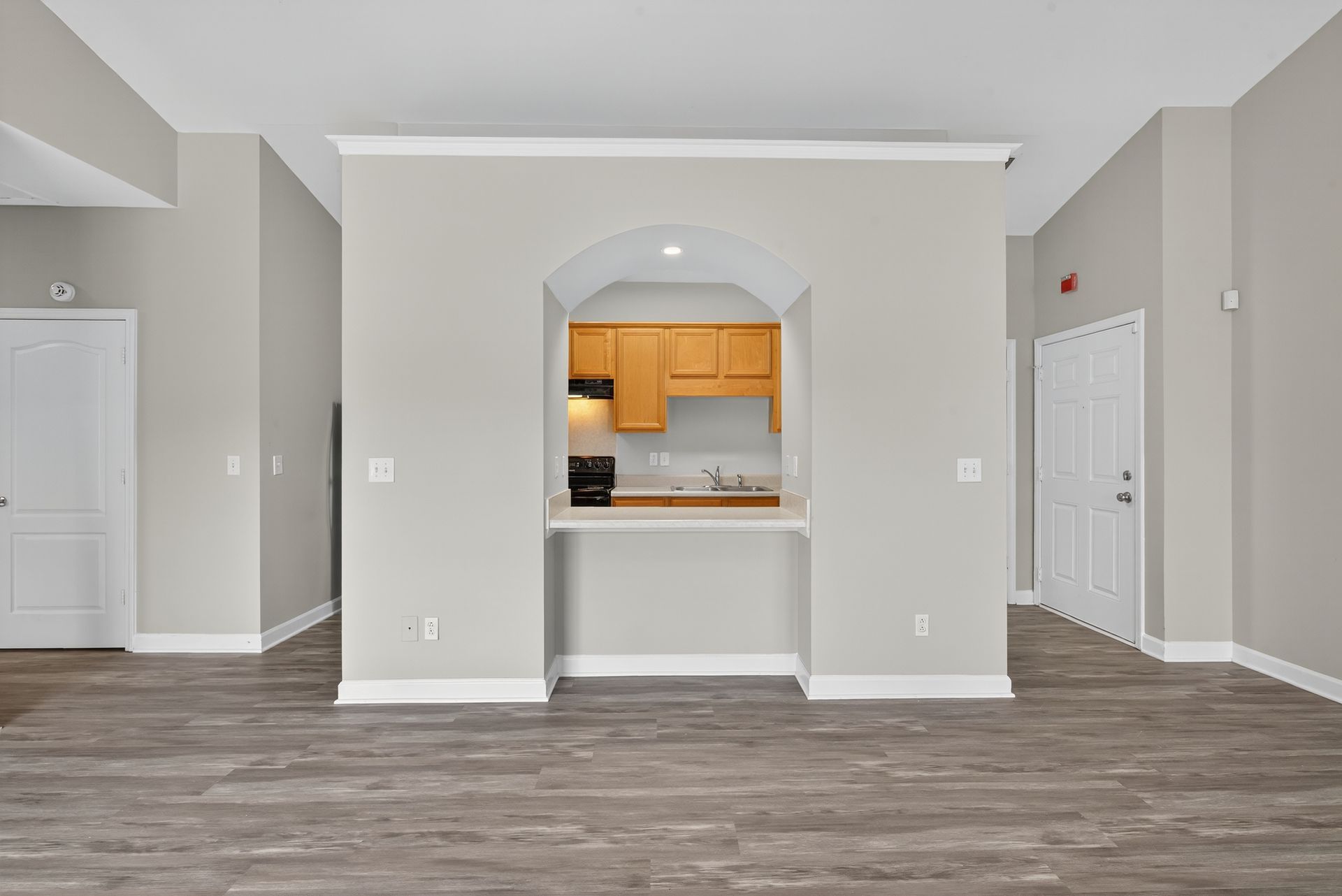 A view of an open-concept room with wood-look flooring, light gray walls, and a central arched pass-through to a kitchen.