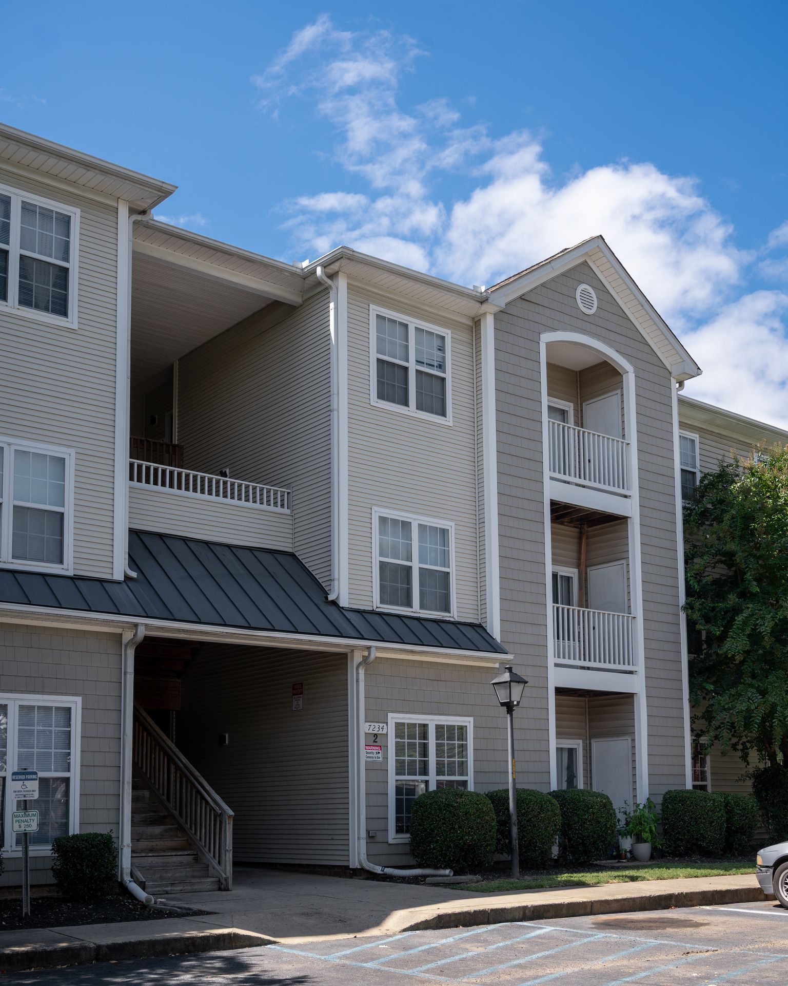 Multi-story apartment building with beige siding, grey trim, balconies, a ground-level breezeway, and blue sky above.