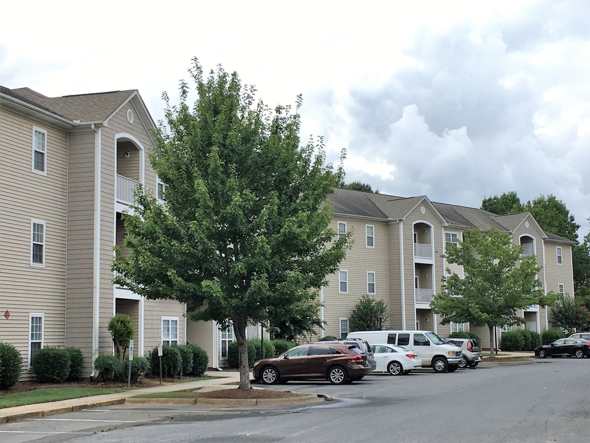 Tan multi-story apartment buildings with parking in front under a cloudy sky.