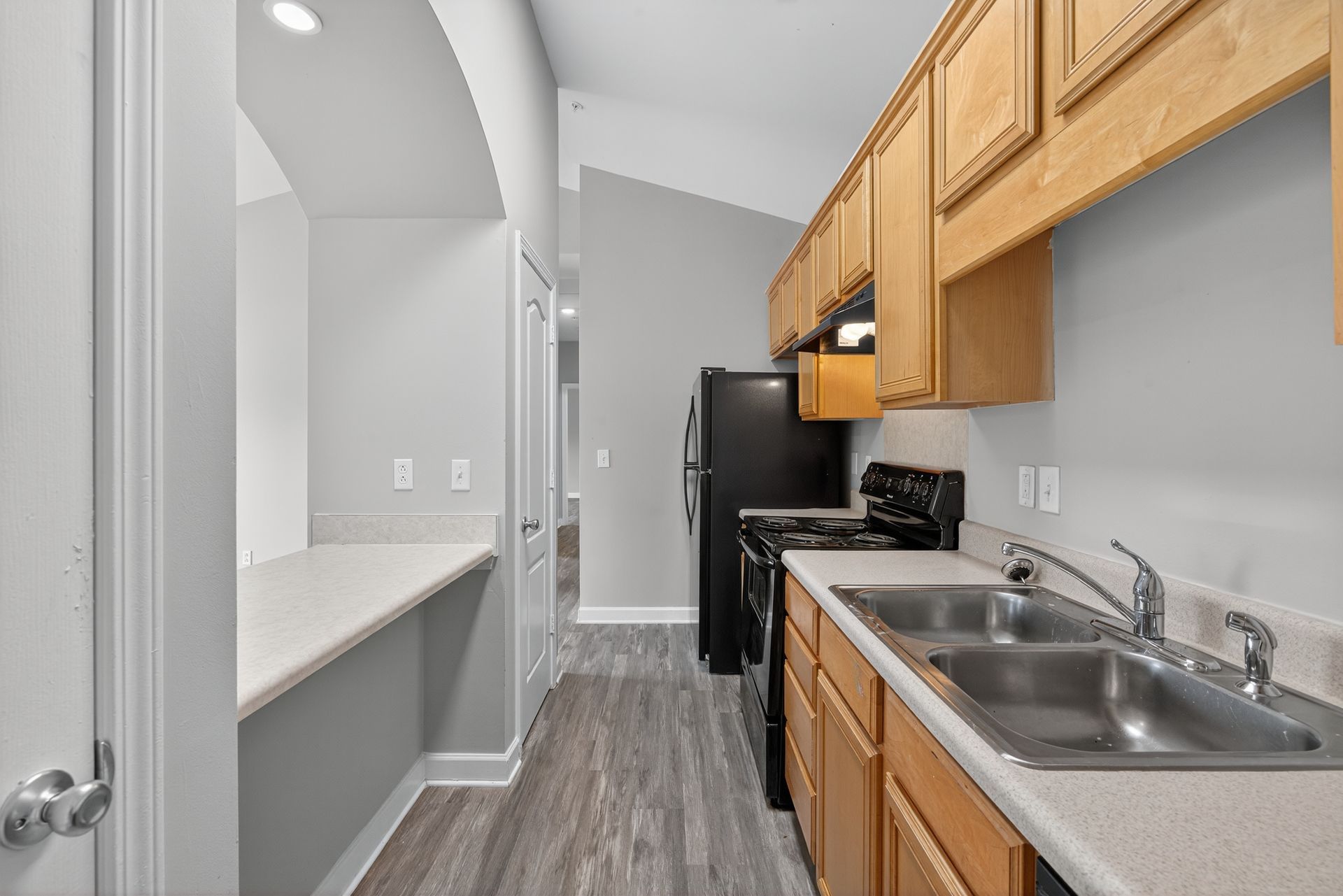 A galley-style kitchen featuring light oak cabinets, gray walls, wood-look flooring, and a black range and refrigerator.