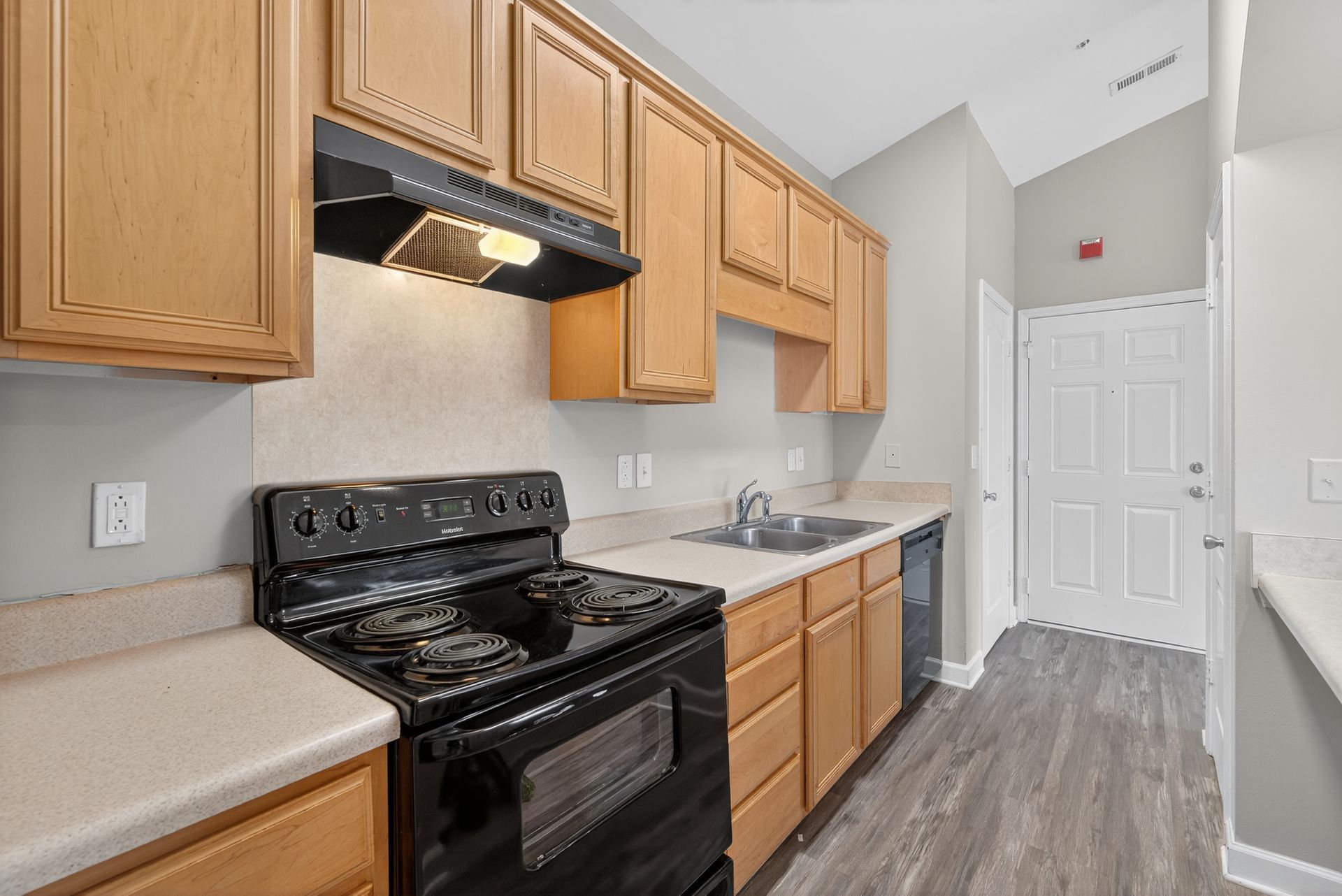 A kitchen with light wood cabinets, black stove, beige countertops, and grey plank flooring leading to a white door.