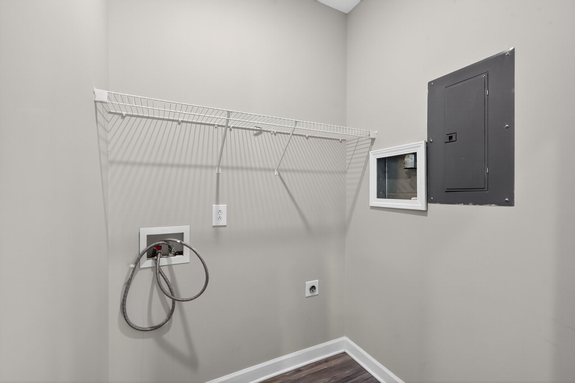An empty laundry area with light gray walls, a metal wire shelf, utility connections, and an electrical breaker panel.