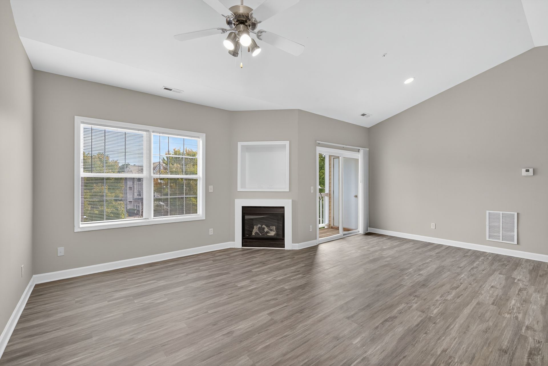 A bright, empty living room with light gray walls, wood-look flooring, a fireplace, ceiling fan, and sliding glass door.