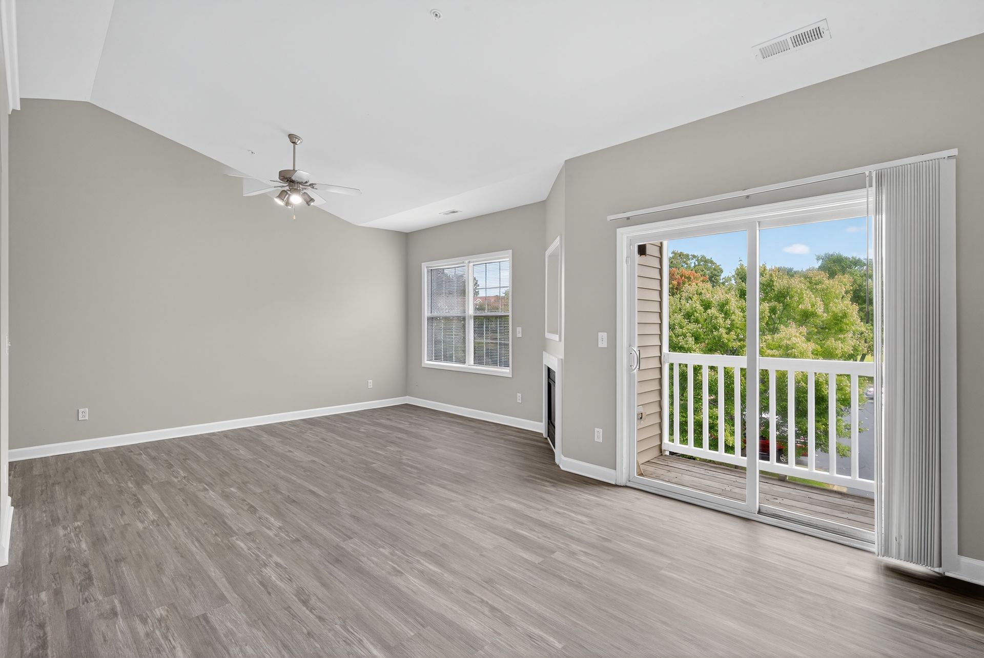 An empty living room with light gray walls, wood-look flooring, a ceiling fan, a window, and a sliding glass door to a deck.