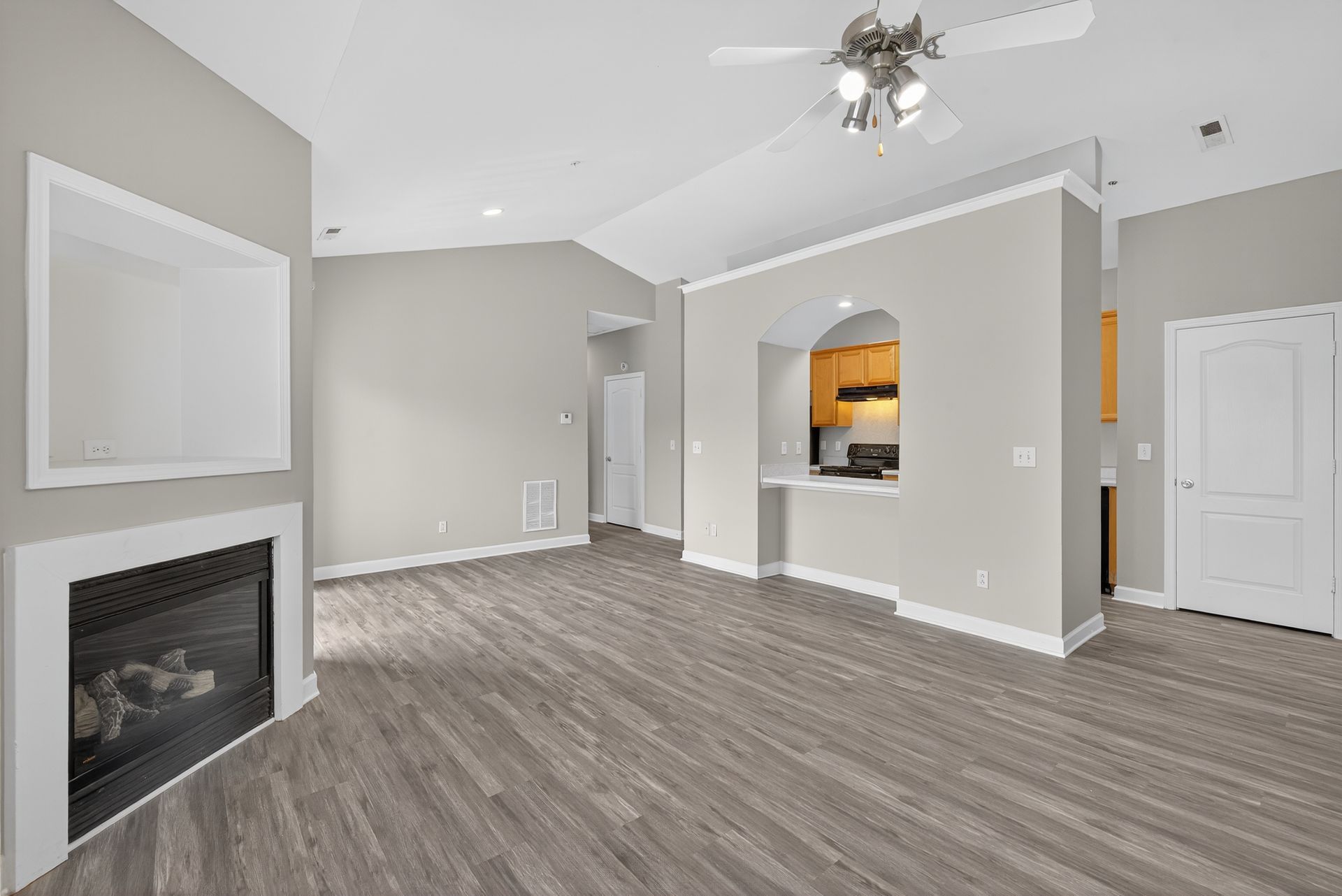 A modern living room with gray wood floors, a fireplace, and an open doorway leading to a kitchen with light wood cabinets.