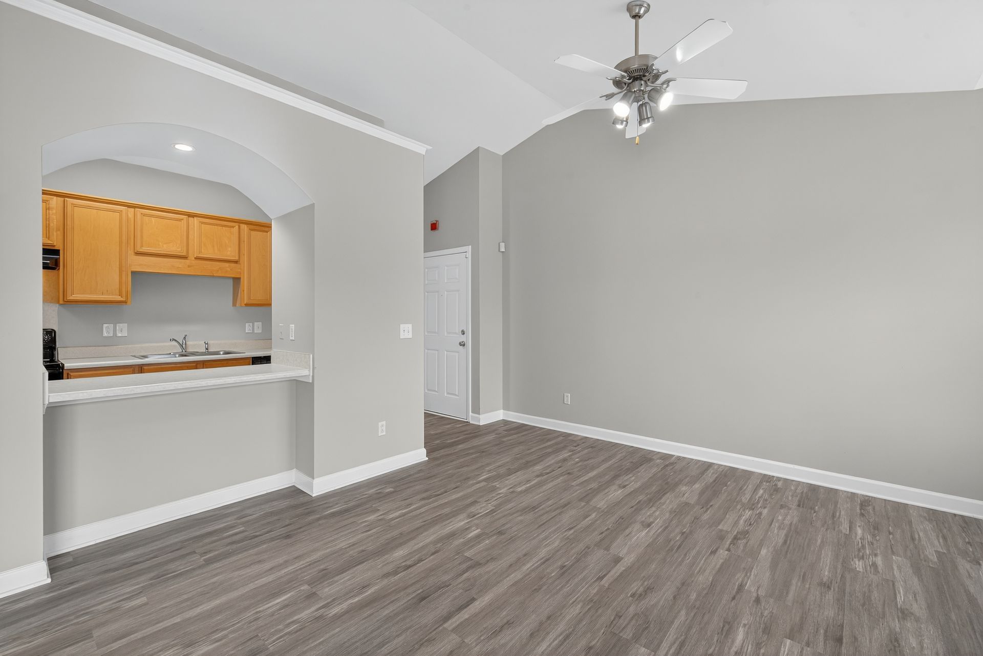 An open-concept living area with grey walls, light-toned wood flooring, a white ceiling fan, and a wood-cabinet kitchenette.
