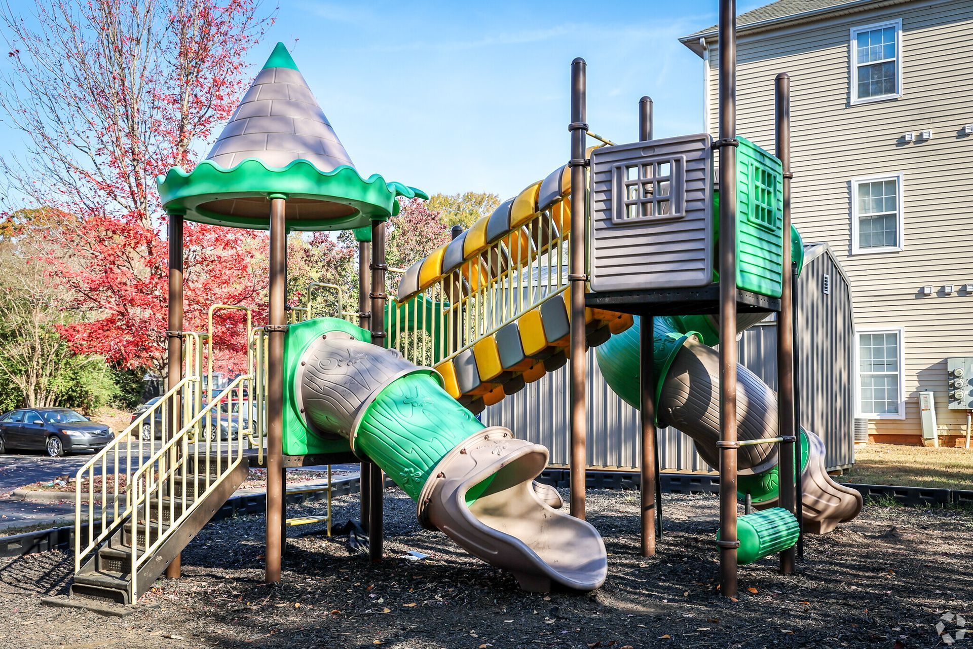 A colorful modular playground structure with slides, stairs, and a roof, set in a yard near a multi-story building.