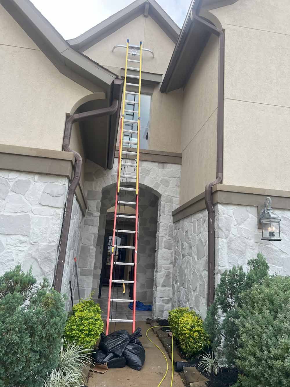 Ladder against a house, reaching to a window. The house has stone and tan siding.