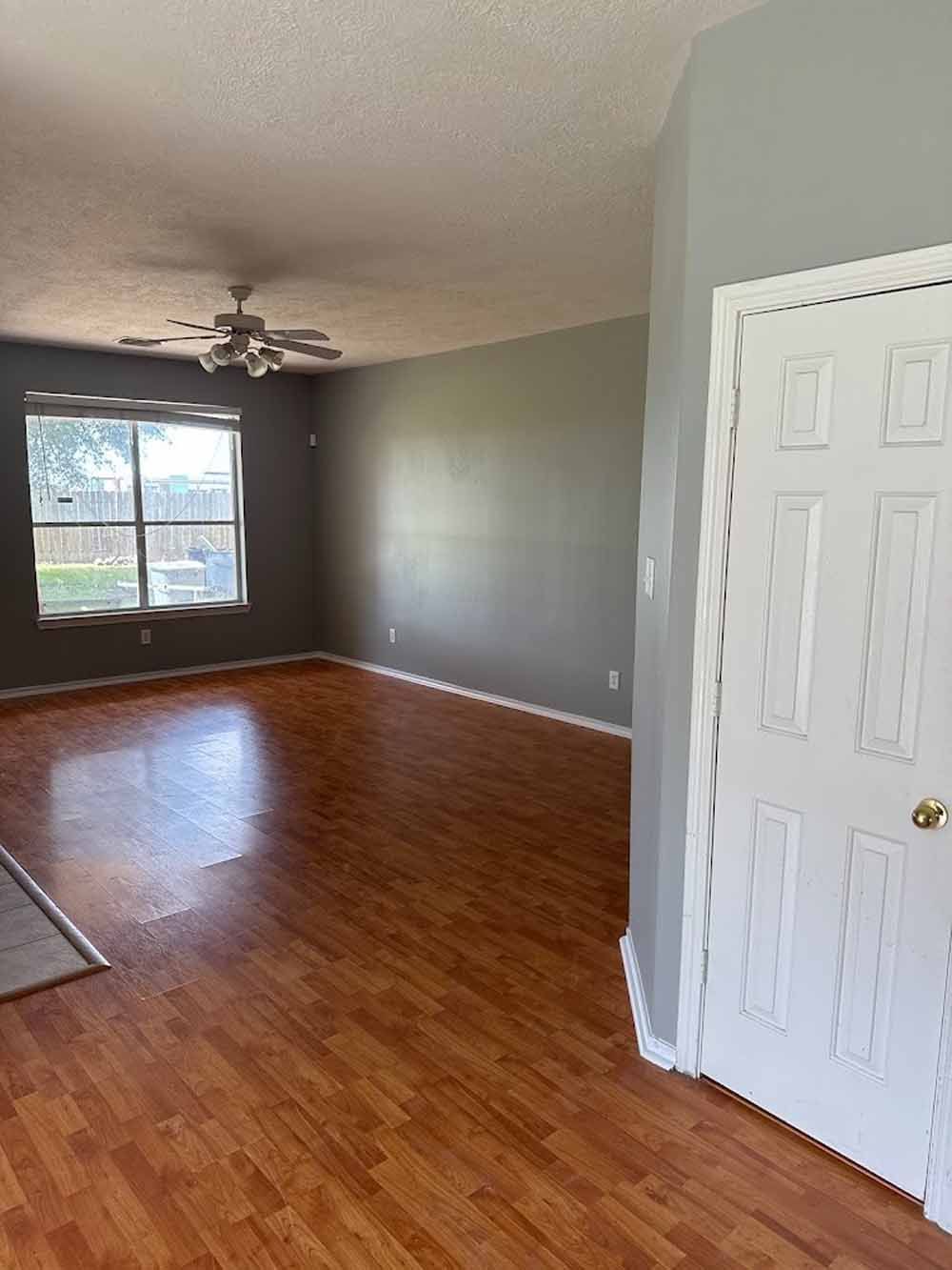 A living room with wood flooring, gray walls, and a white door. There is a window and ceiling fan.