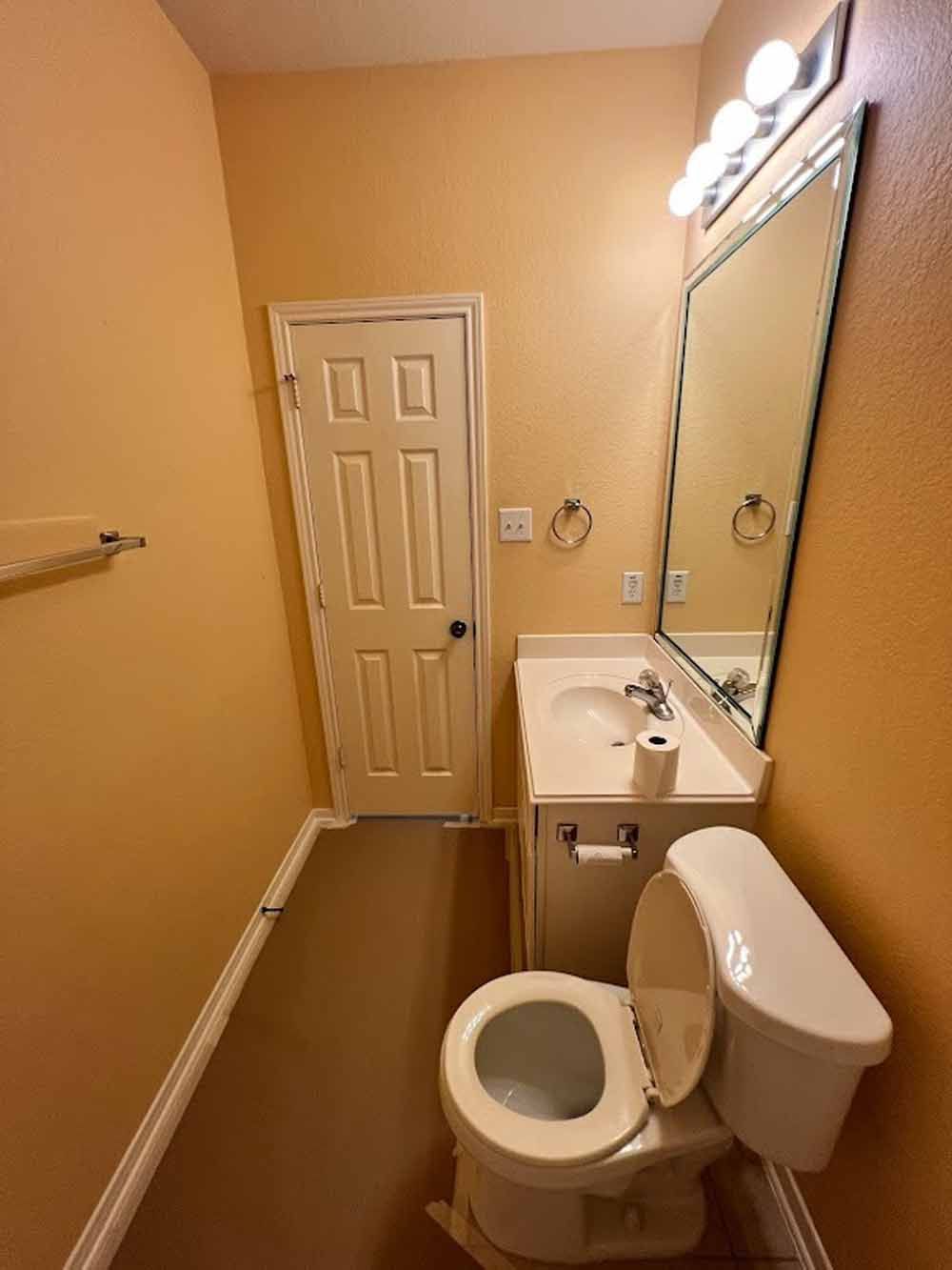 A small bathroom with a toilet, sink, mirror, and closed door, all under warm lighting.