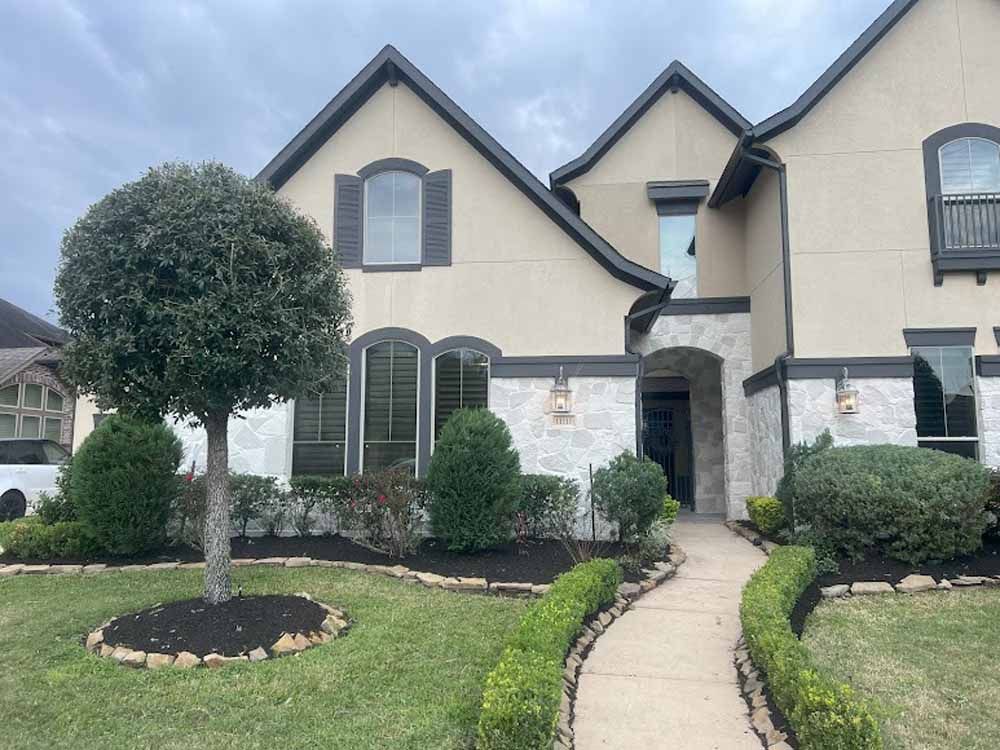 Beige two-story house with dark trim and stone accents, neatly landscaped front yard, cloudy sky.