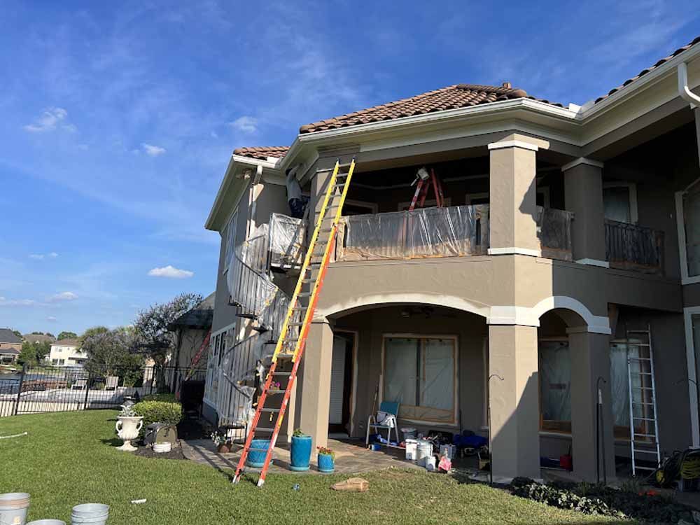 Two-story house being painted; orange ladder up to the balcony. Green grass, blue sky.
