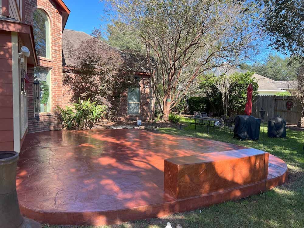 Red-stained concrete patio with a built-in bench, next to a brick house and a grassy yard.