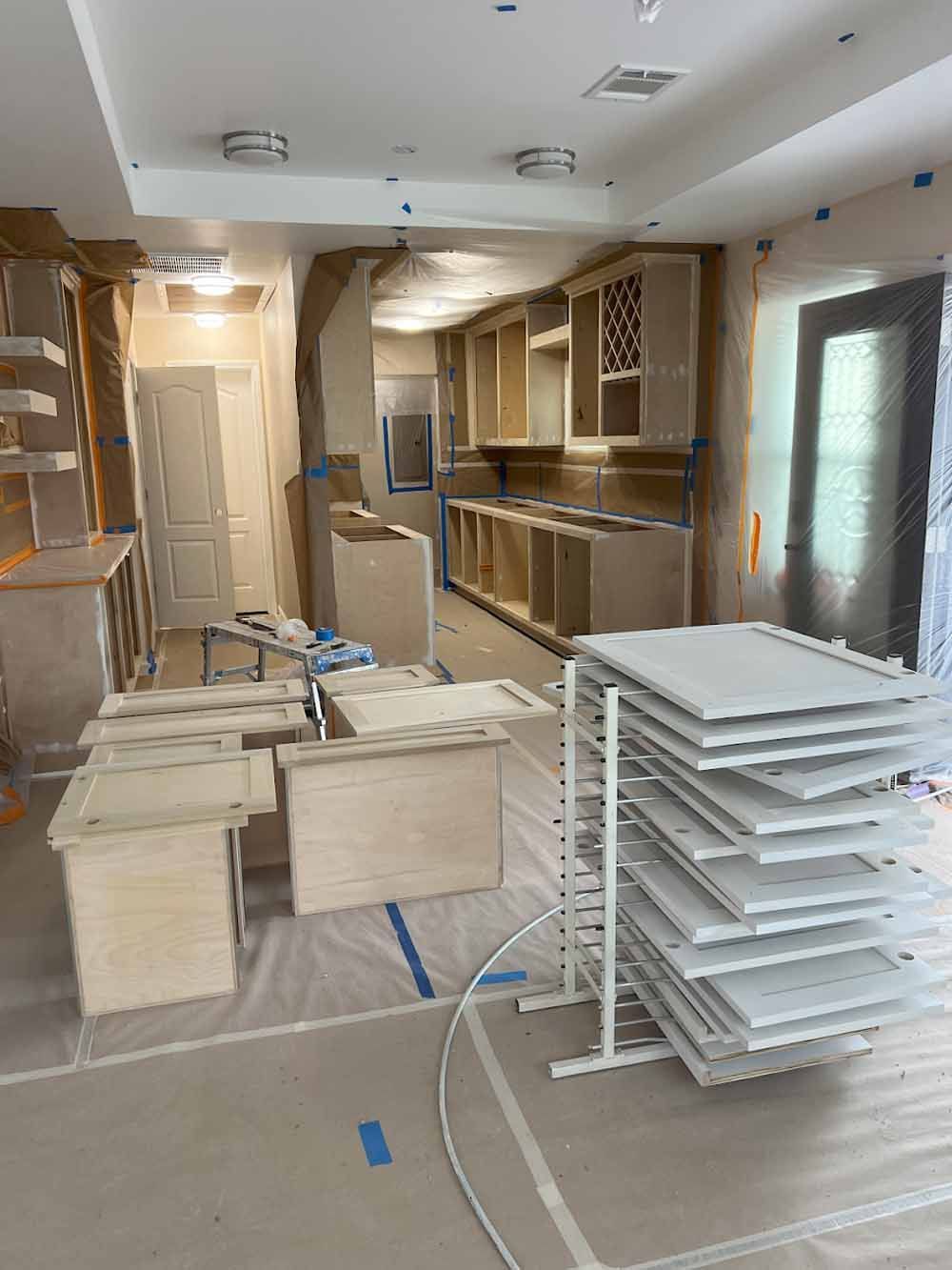 Kitchen cabinets under construction; doors drying on rack. Room covered in protective sheeting.