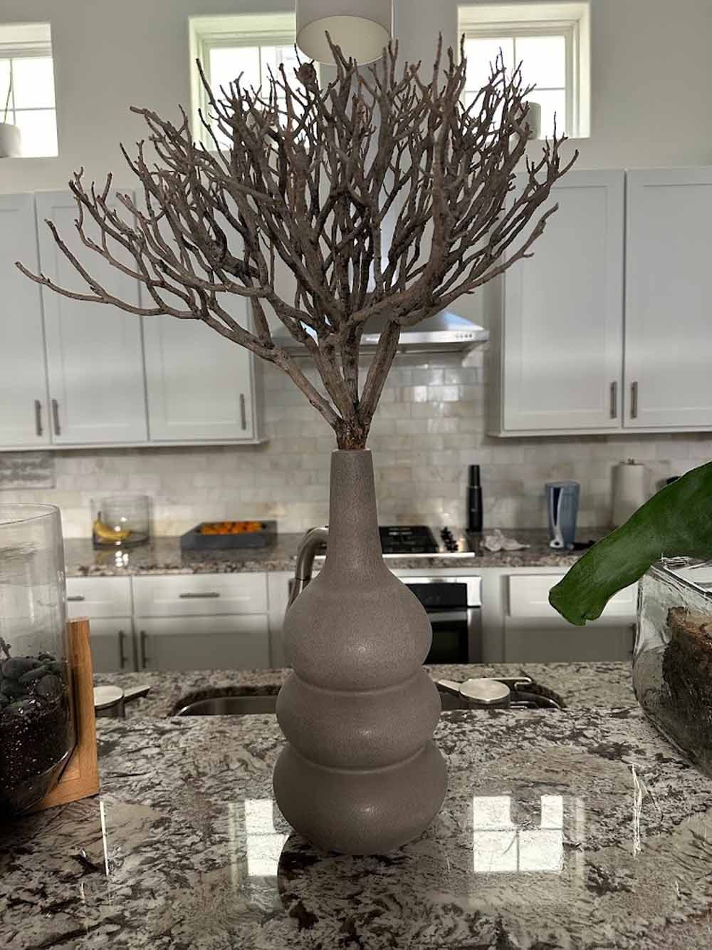 Tall gray vase with brown branches on a granite countertop in a kitchen.