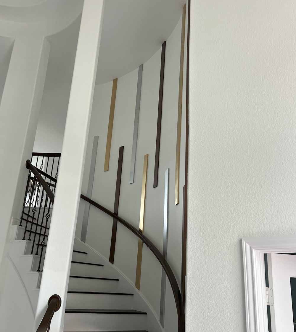 Stairwell with white walls, curved banister, and vertical metallic decorative strips in golds, silvers, and browns.