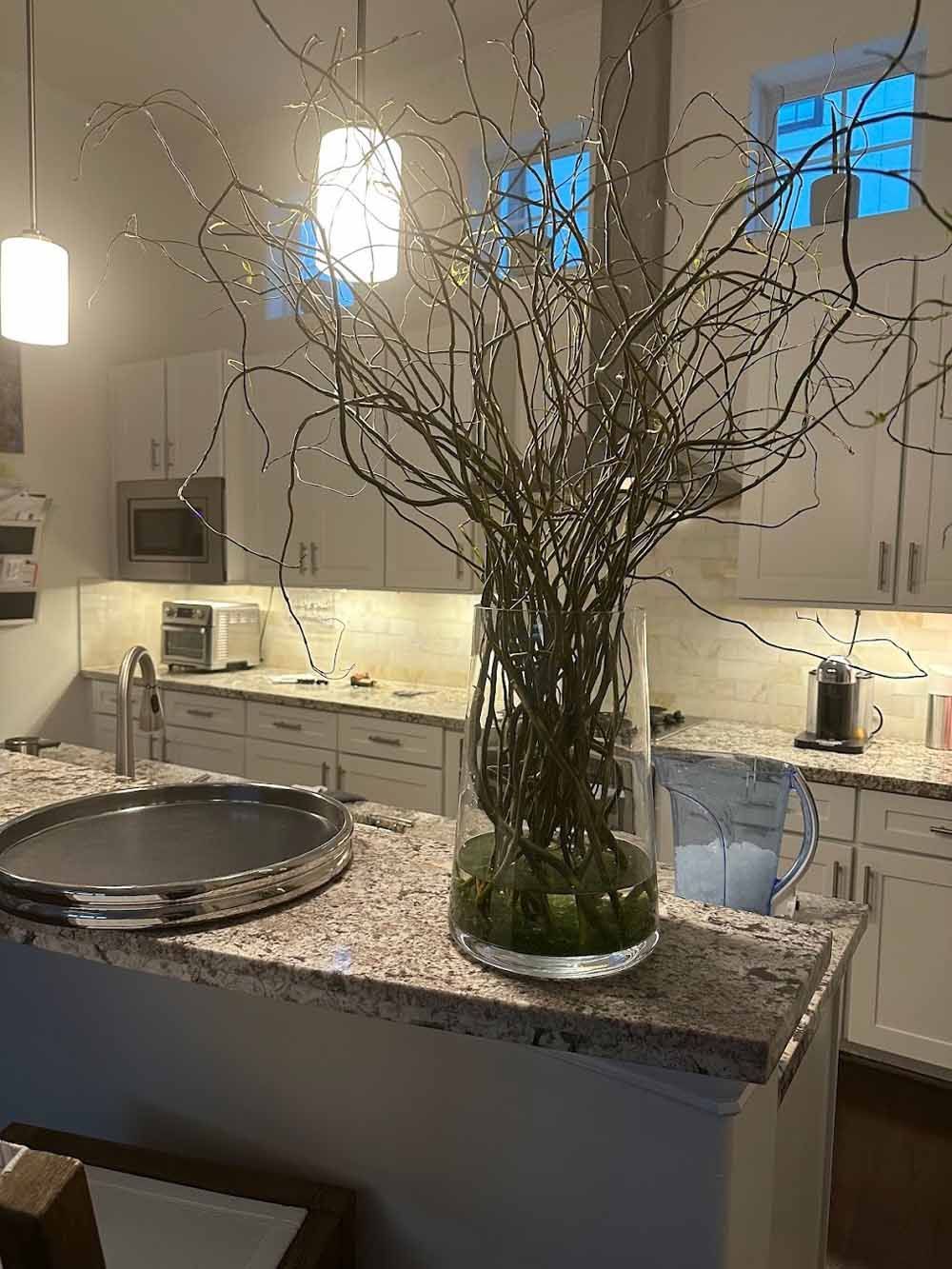 Kitchen with a large glass vase of branches on a countertop. White cabinets, island, and appliances are in view.