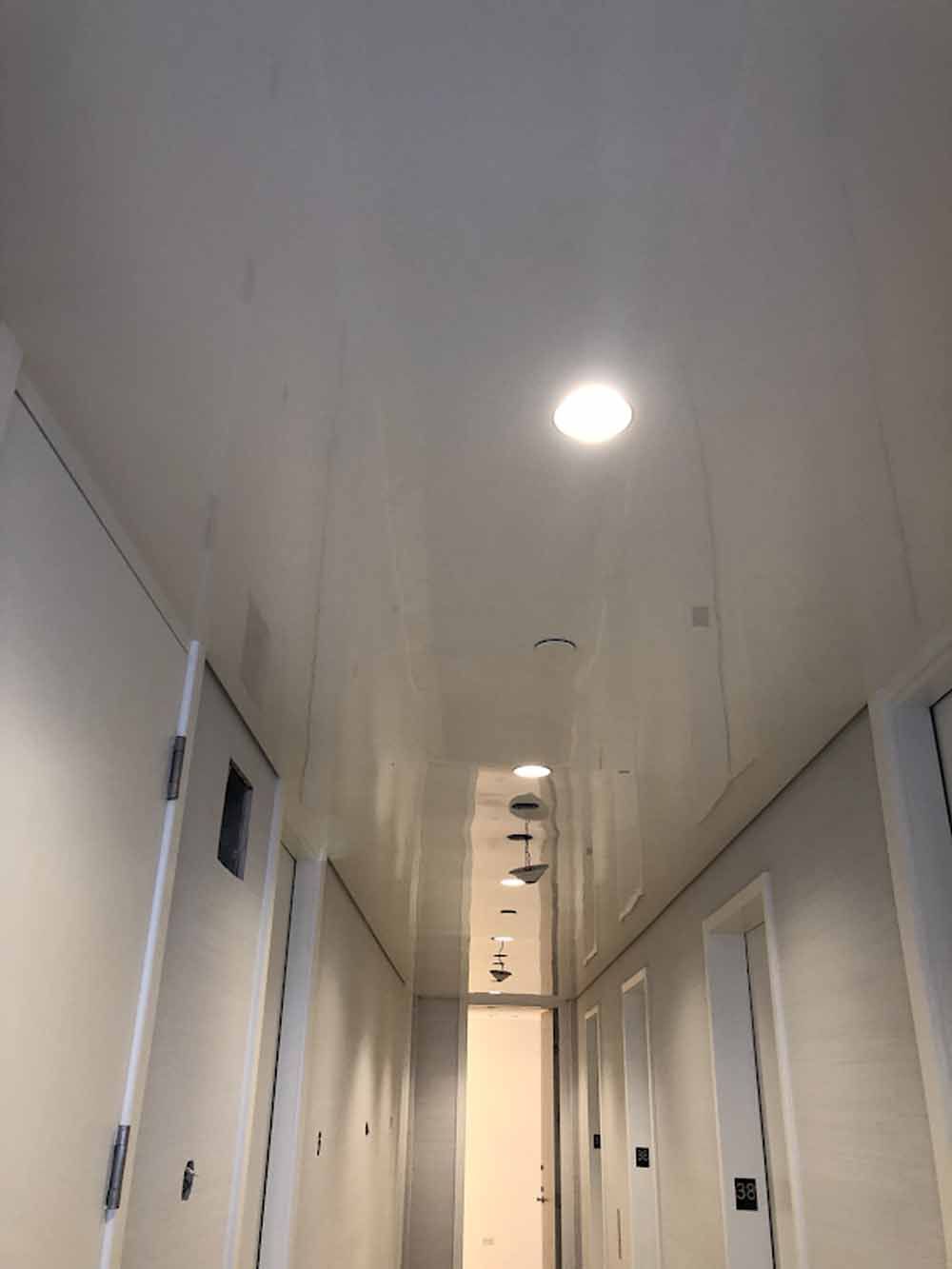 Narrow hallway with white walls and ceiling, several doors on either side, bright overhead lights.