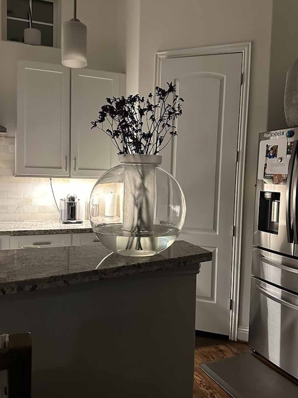 Kitchen island with globe vase of dark flowers, stainless steel refrigerator, white cabinets, and granite countertop.