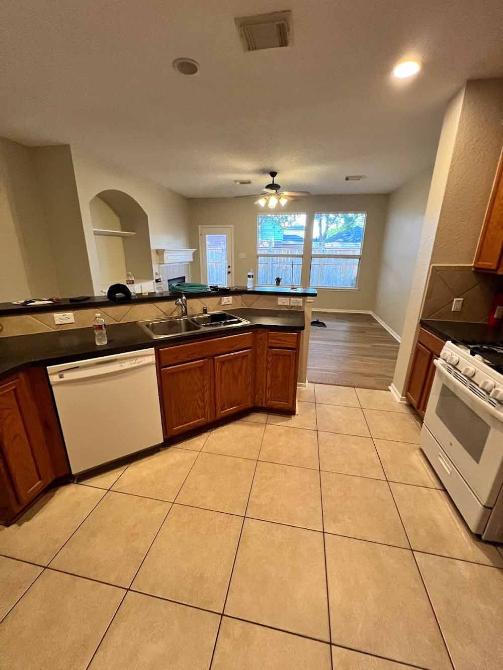 Kitchen with brown cabinets, white appliances, and tan tile floor, leading to a living area with windows.