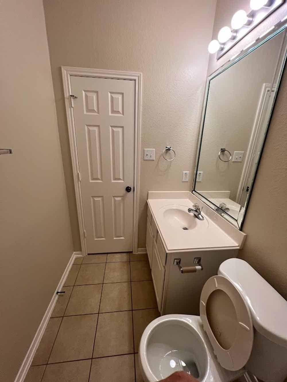 Bathroom with a white door, toilet, sink, and mirror. The floor is tiled.