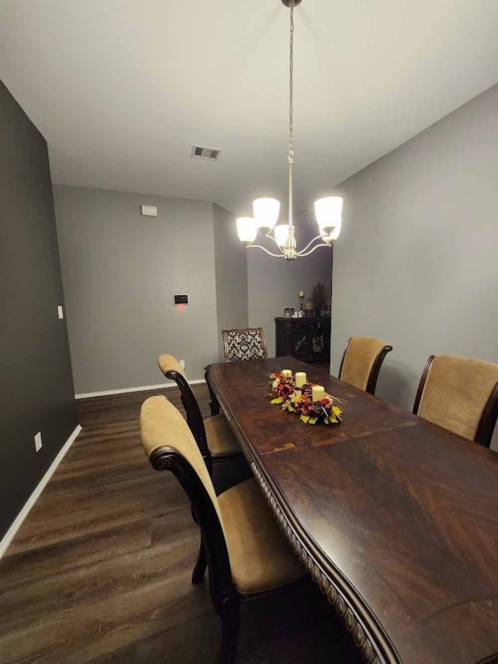 Dining room with long wooden table, six chairs, chandelier, dark walls, and a centerpiece.