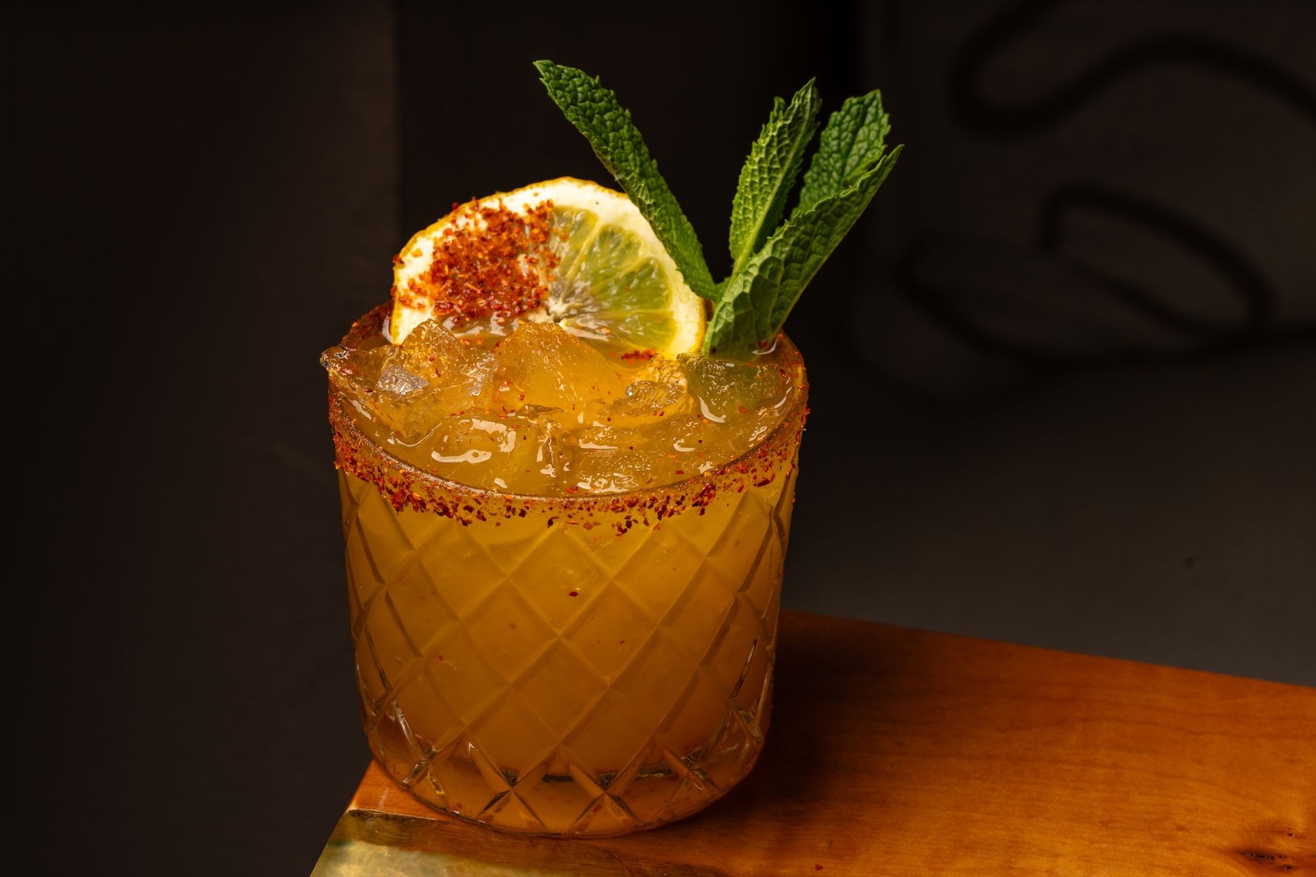 Orange colored cocktail with tajin rim, lemon garnish and mint.