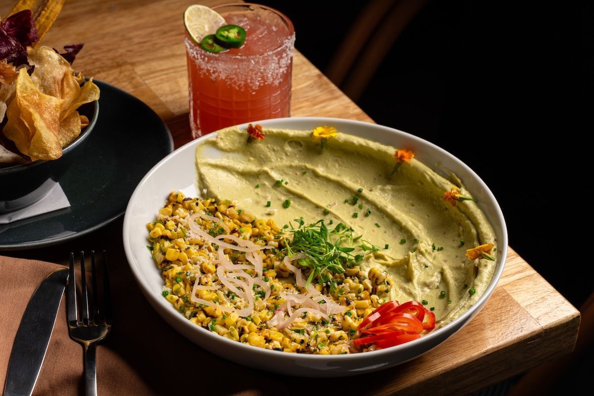 Brisa street corn and guacamole appetizer with pink cocktail and side of chips, sitting on the edge of wood table 