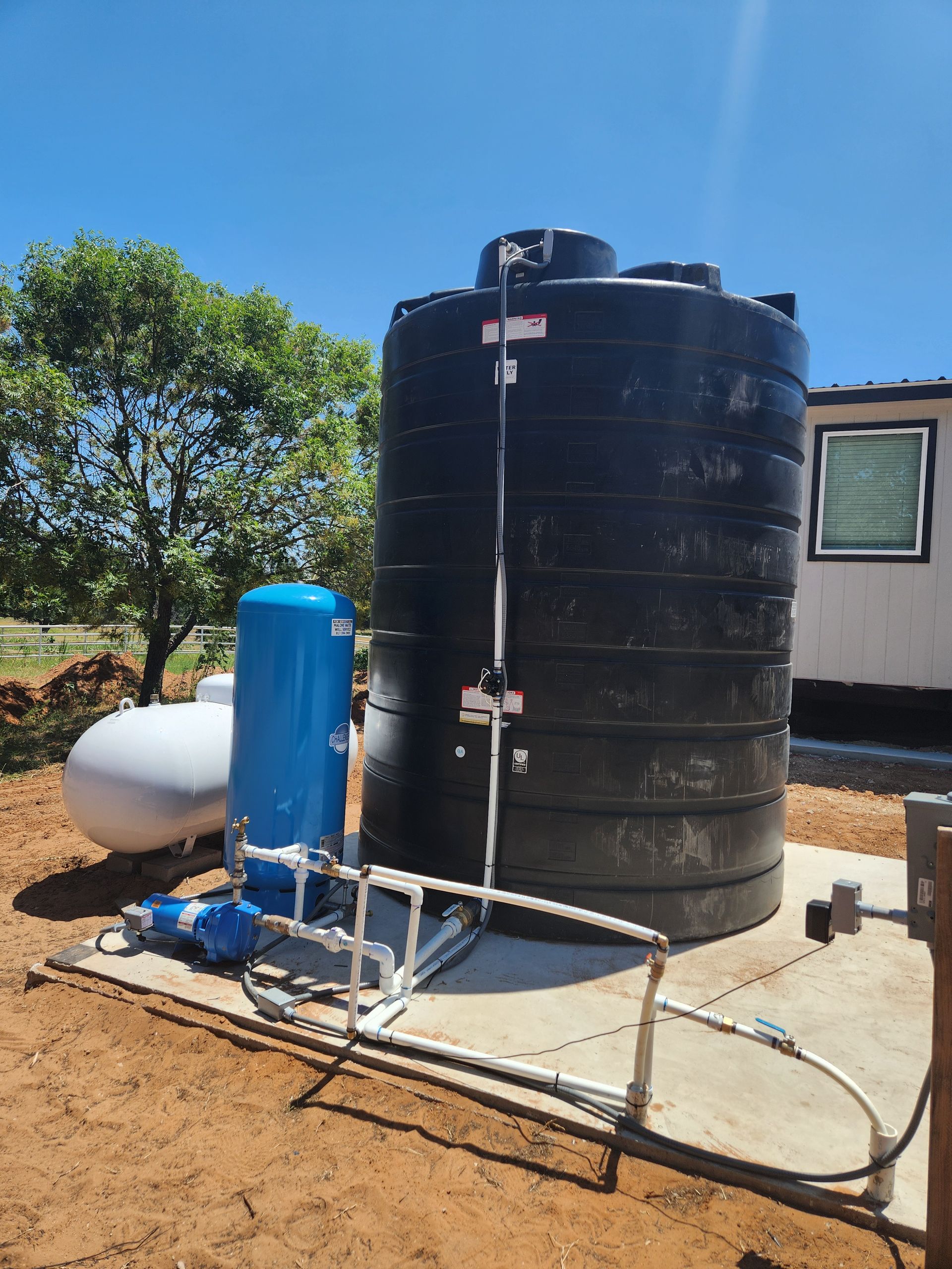 A large black water tank is sitting in the dirt next to a blue pump.
