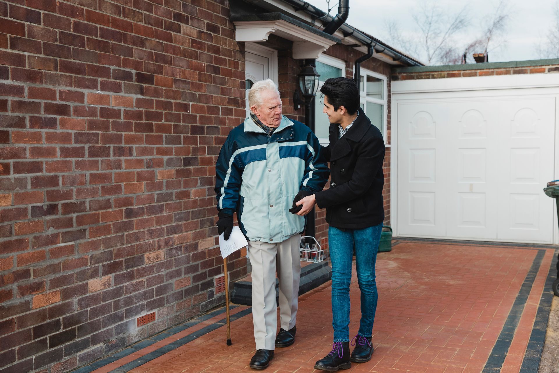 A young man is helping an older man walk in front of a brick building.
