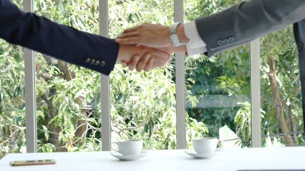 Two businessmen are shaking hands over a table with cups of coffee.