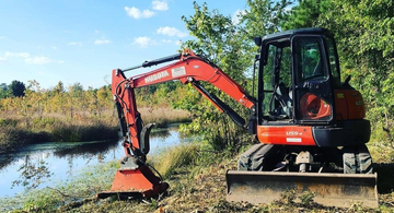 A red and black excavator is sitting next to a body of water.