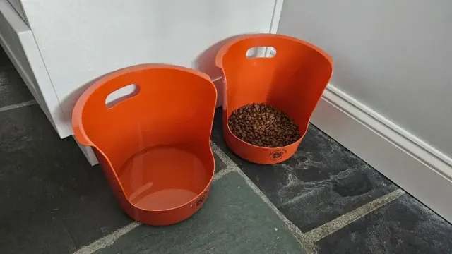 Two orange pet feeding buckets on a dark tiled floor, one with dry kibble inside.