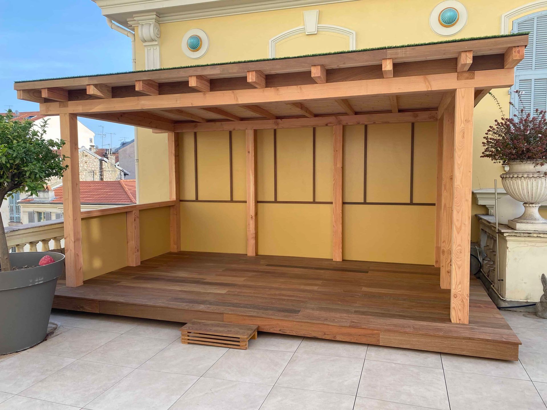 Une pergola en bois est installée sur une terrasse devant un bâtiment jaune.