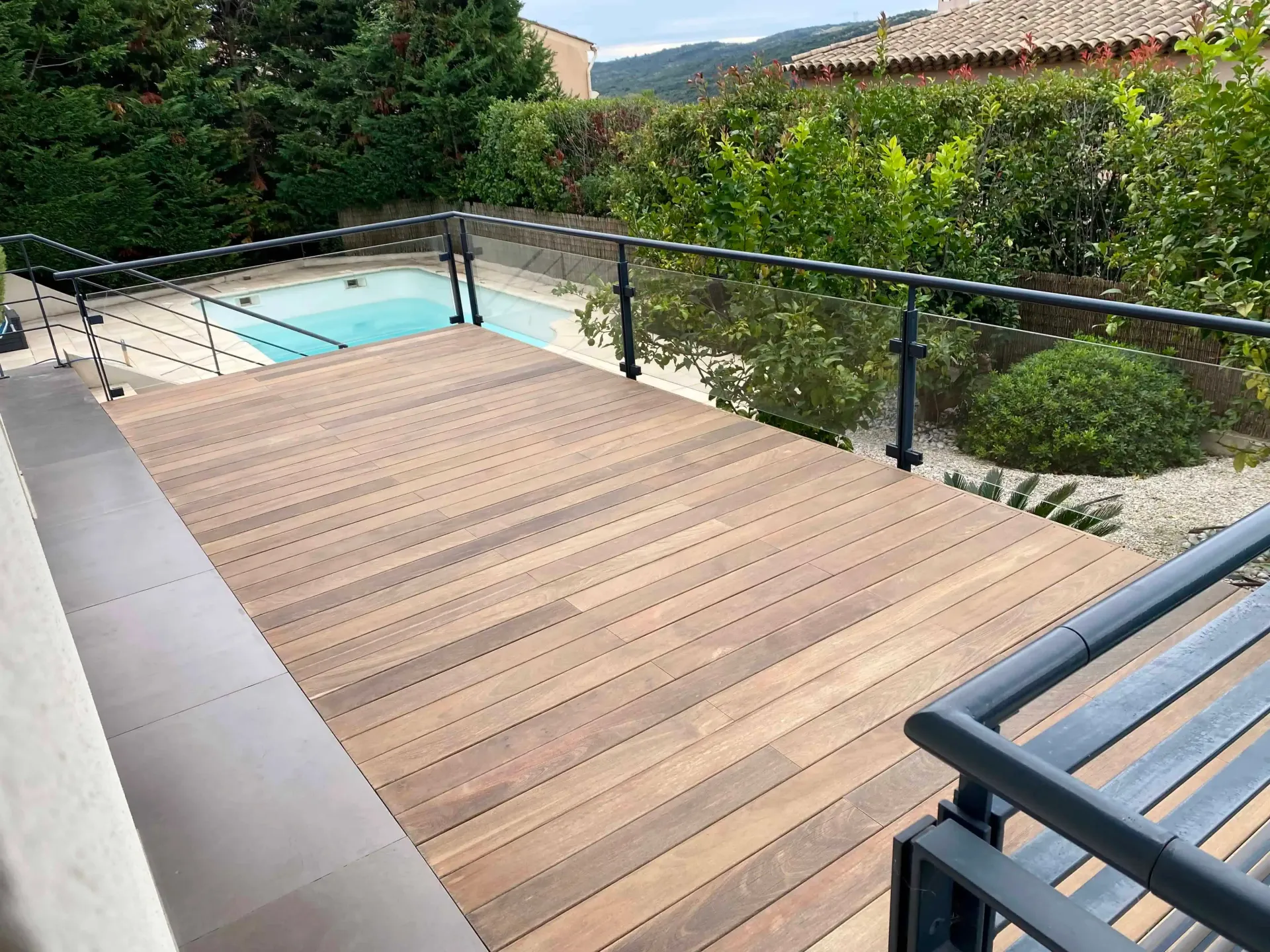 Terrasse en bois rénovée avec balustrade noire donnant sur une piscine et un jardin.