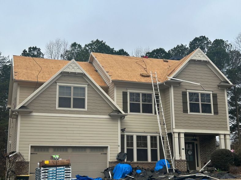 Two-story house under construction with exposed roof sheathing, ladders, and blue tarps in the front yard