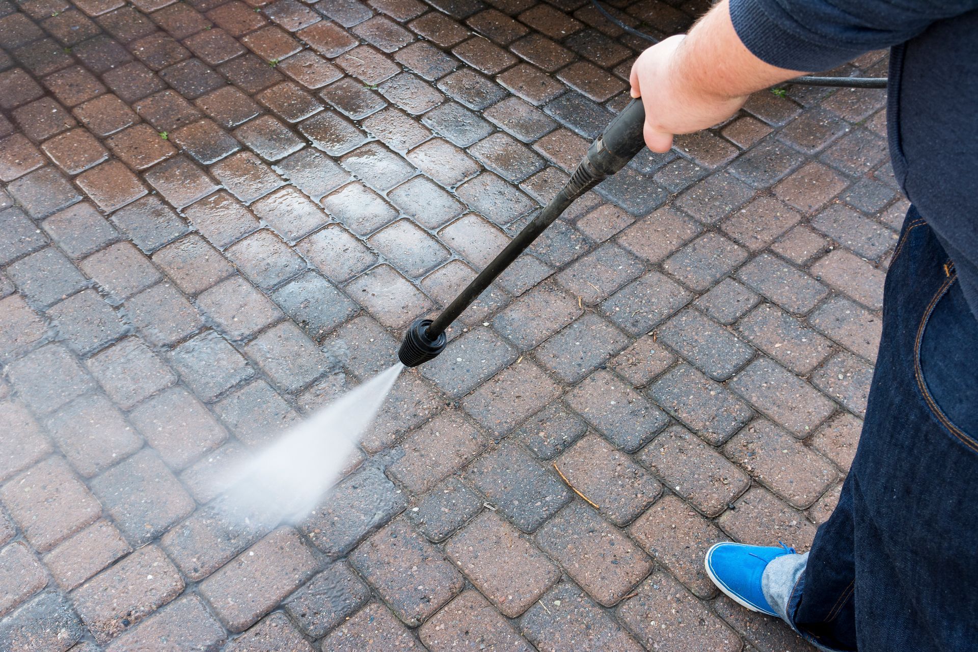 A person is using a high pressure washer to clean a brick driveway.