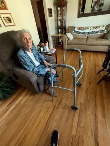 Elderly person sitting in chair, walker nearby. In a living room setting, hardwood floors, sofa in background.
