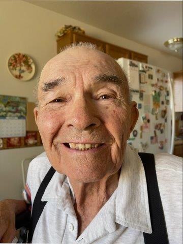 Smiling elderly man with suspenders, seated indoors. Kitchen in background.