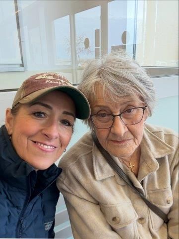Woman smiles for selfie with older woman in glasses, indoors.